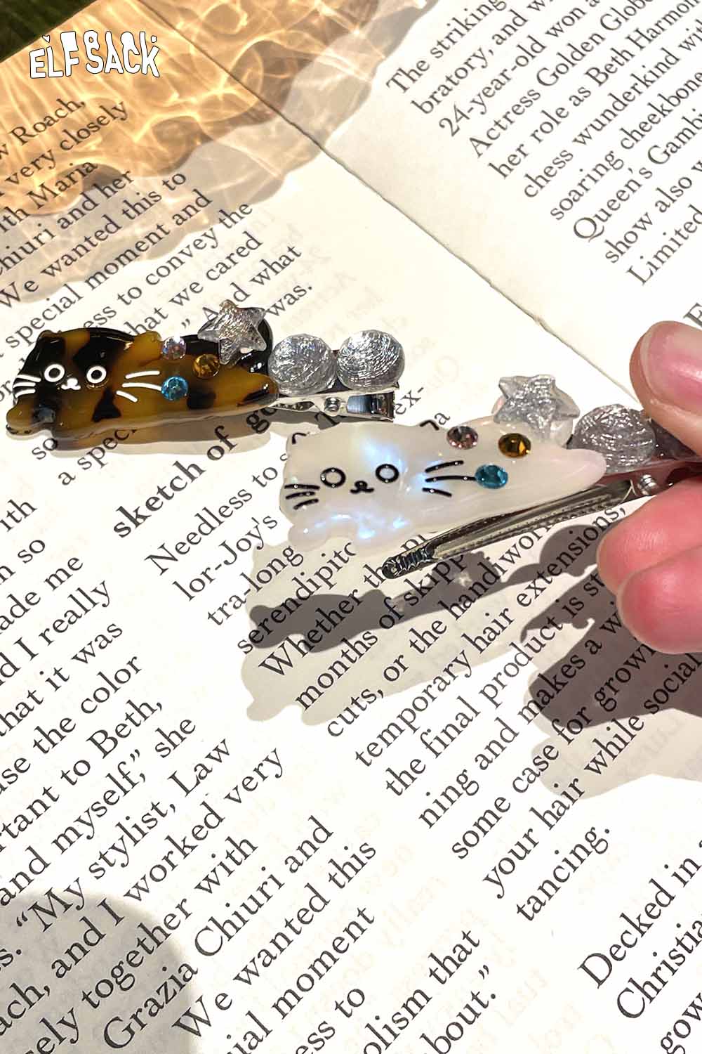 Two ELFSACK acetate kitten hairpins with colorful embellishments on a book page