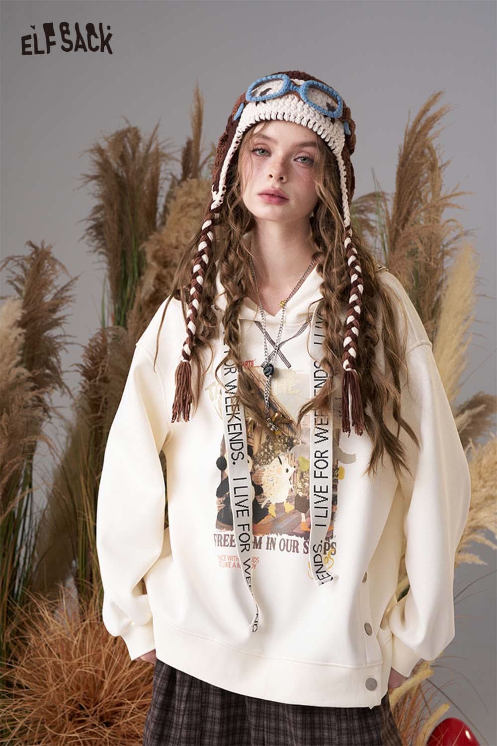 Color blocked crocheted ear flap beanie with braided tassels, worn by a model in a cozy setting