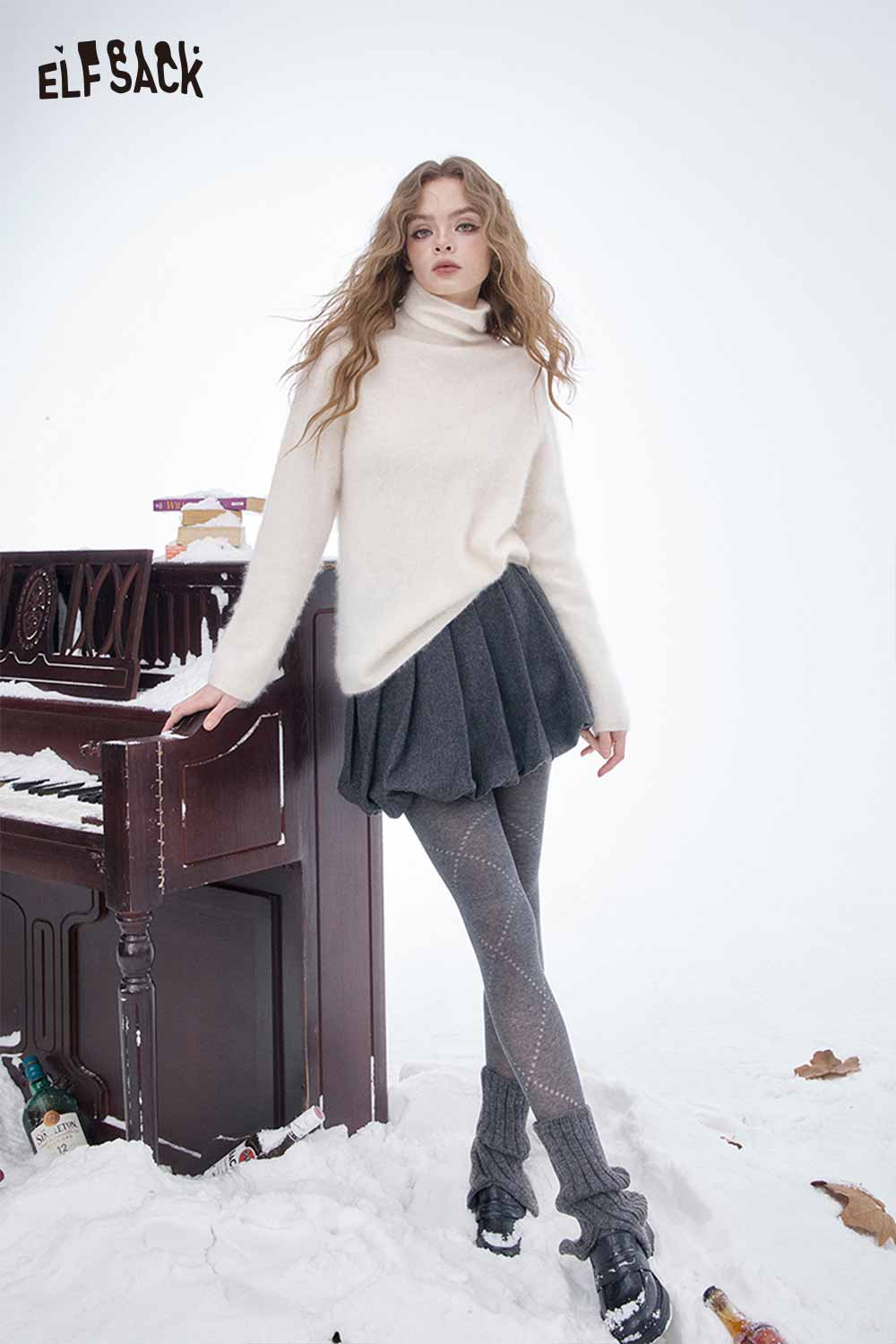 Model wearing a dark gray pleated half skirt paired with a cozy sweater in a snowy setting