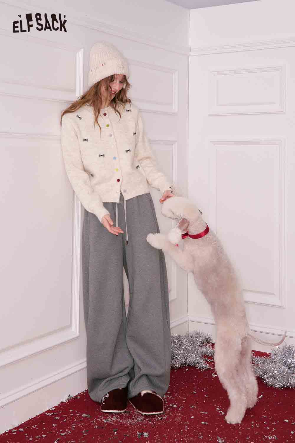 ELFSACK girly bow cardigan in cream color, worn with cozy gray pants and a playful dog