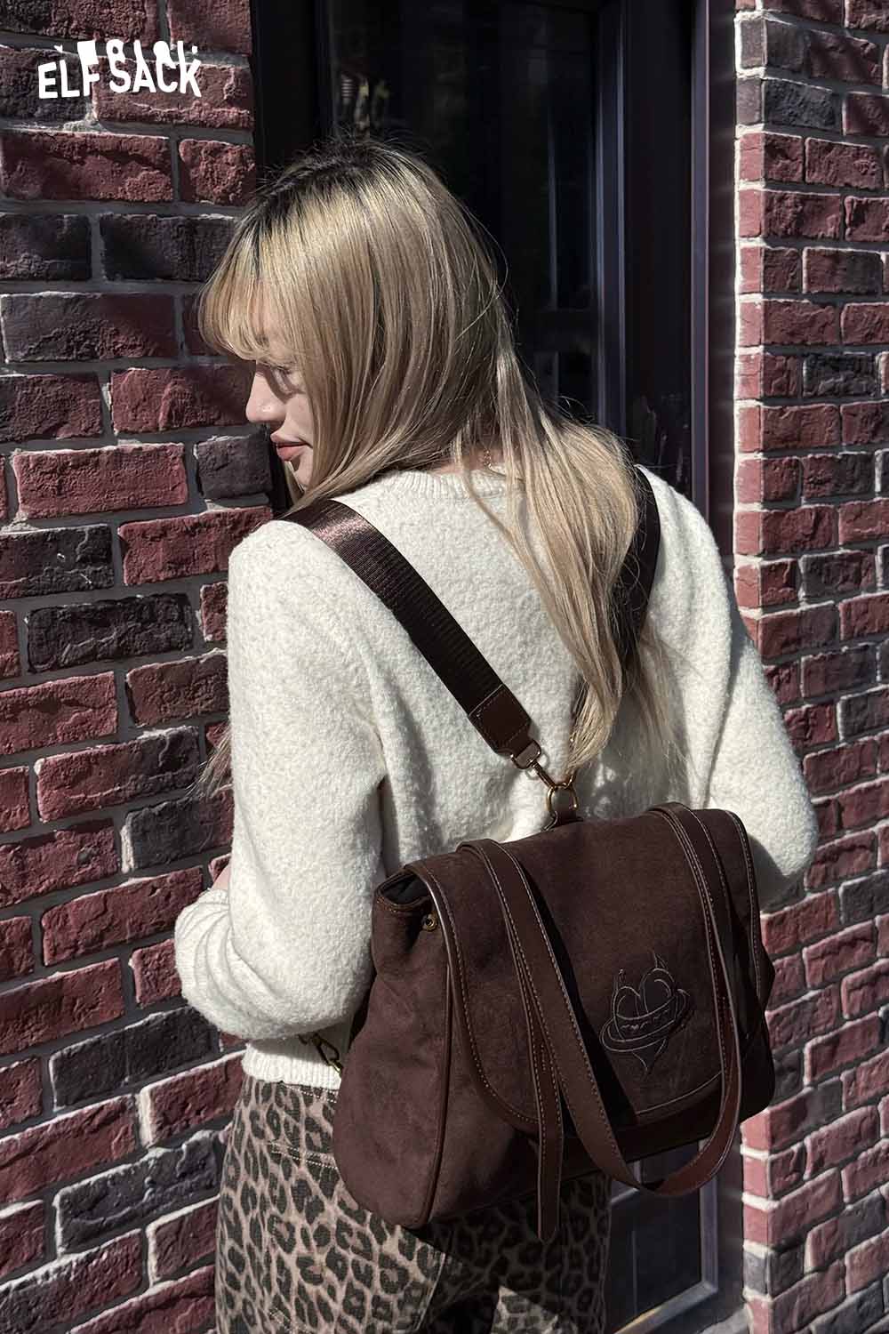 Stylish ELFSACK preppy suede backpack for women, worn with a cozy sweater against a brick wall