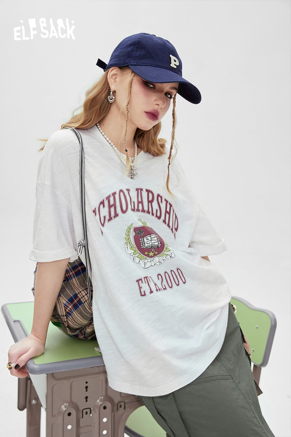 ELFSACK white oversized t-shirt with graphic print and vintage scholarship design worn with green pants and navy cap