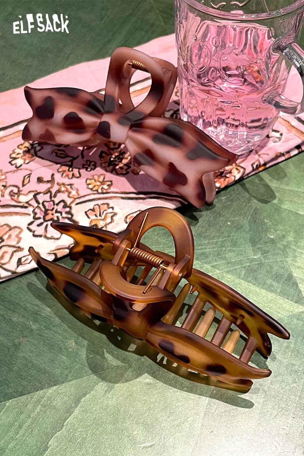 ELFSACK tortoiseshell bow hair claw set on a floral-patterned tablecloth next to a pink glass