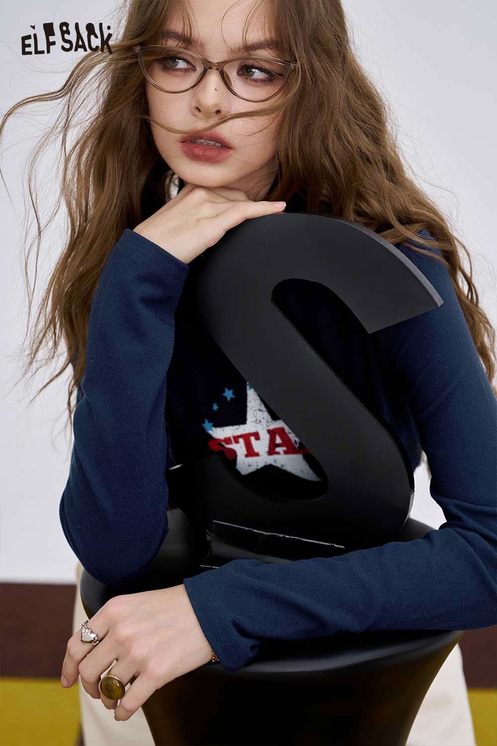 ELFSACK women's graphic long sleeve t-shirt displayed on a model with stylish glasses against a striking backdrop