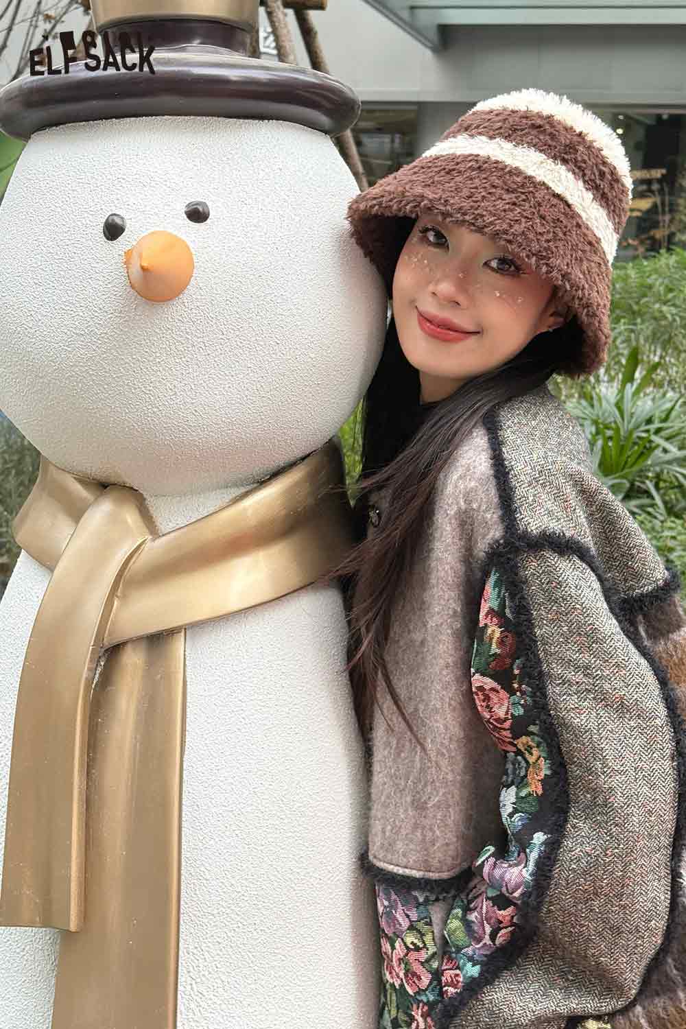ELFSACK women's plush bucket hat in brown with stripes, styled with winter apparel next to a snowman decoration
