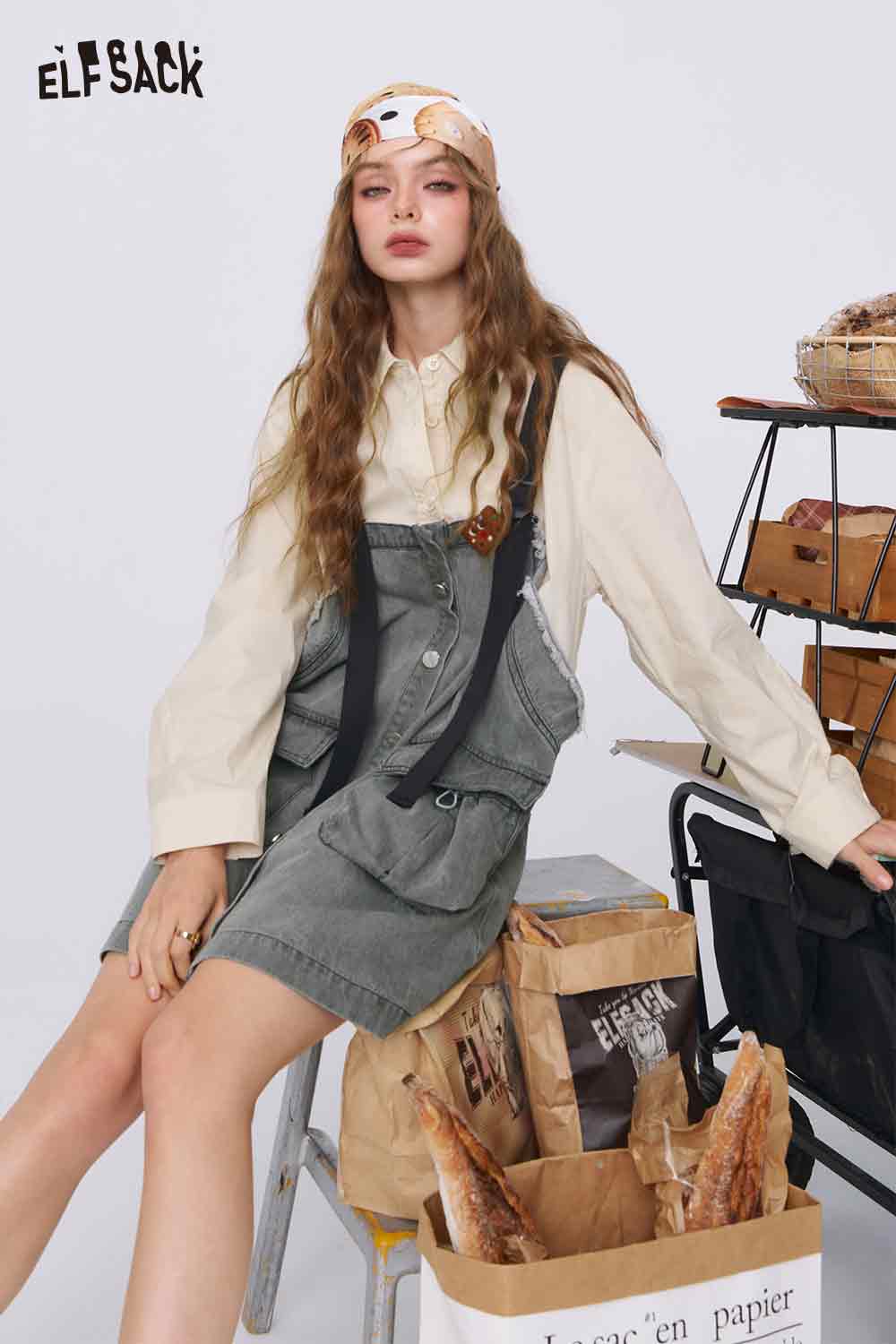 ELFSACK model sitting wearing a denim color-block midi dress pinafore over cream blouse with headscarf and baguettes