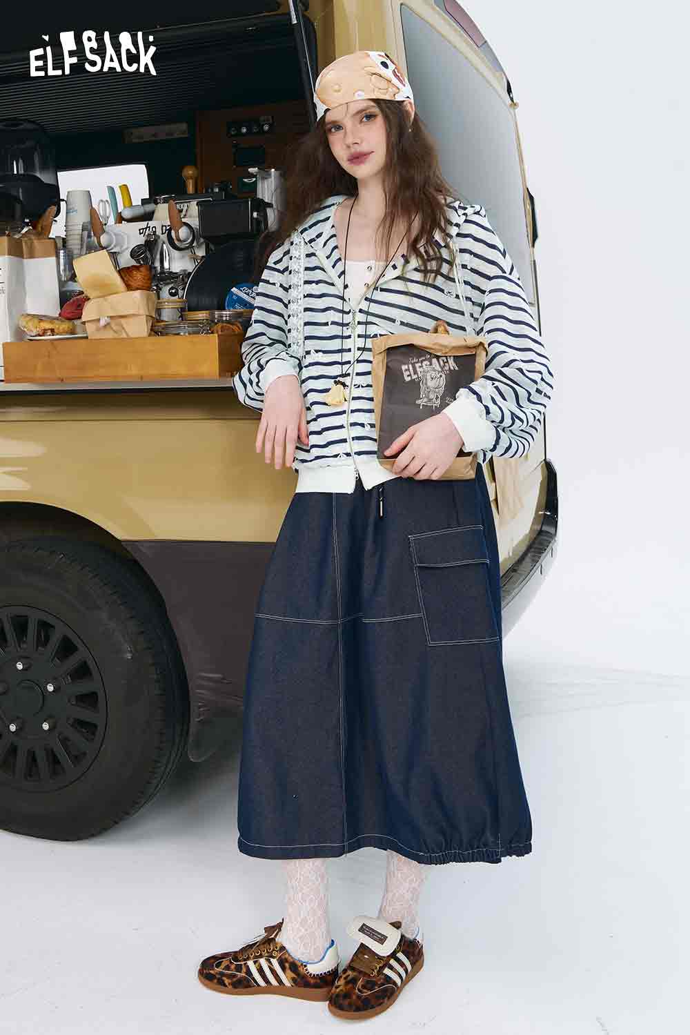 ELFSACK model wearing oversized striped cardigan for women in white navy with denim skirt holding book by coffee van