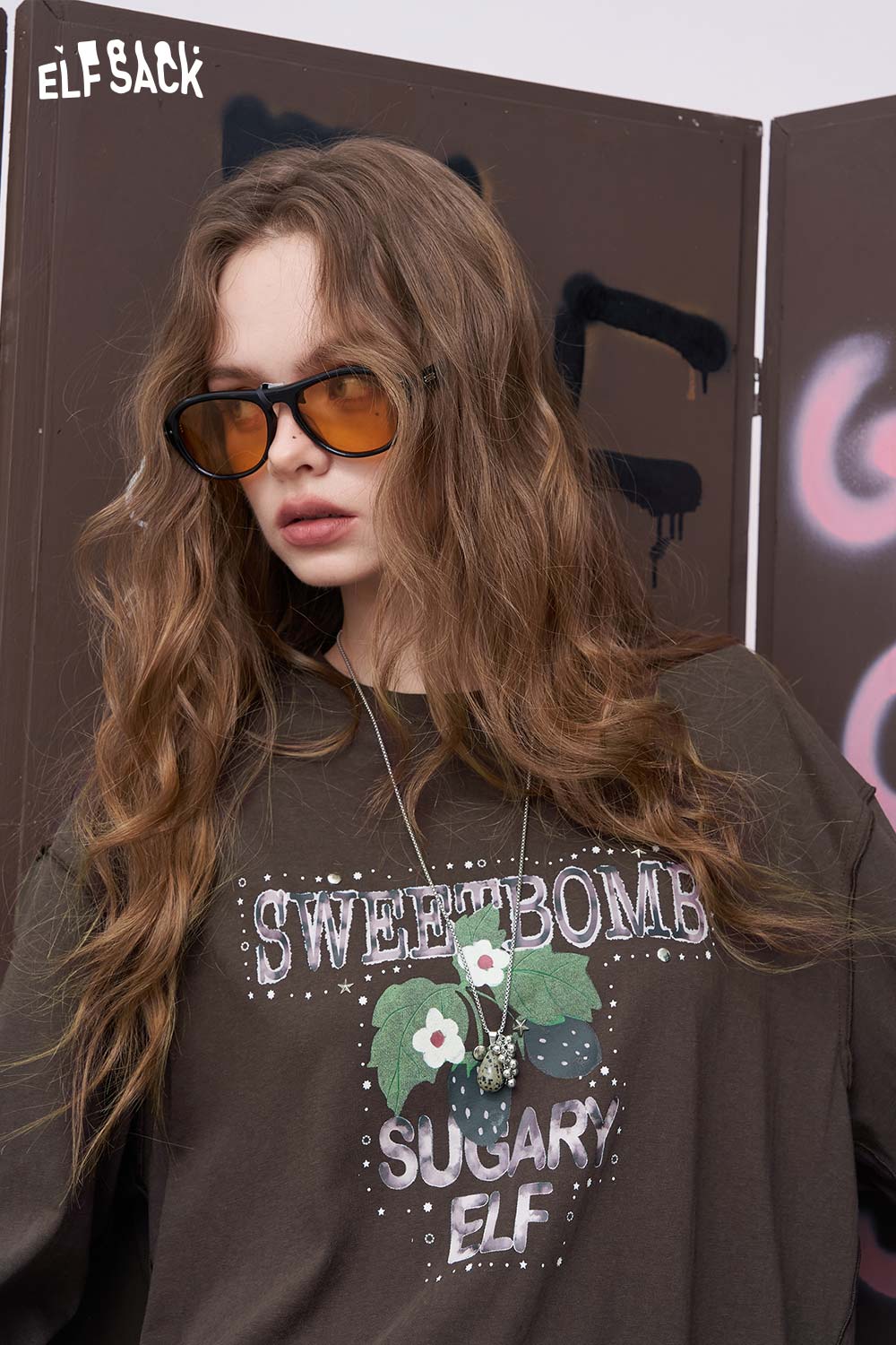 
                  
                    ELFSACK model wearing oversized daisy graphic tee for women in brown with Sweet Bomb floral print and sunglasses
                  
                