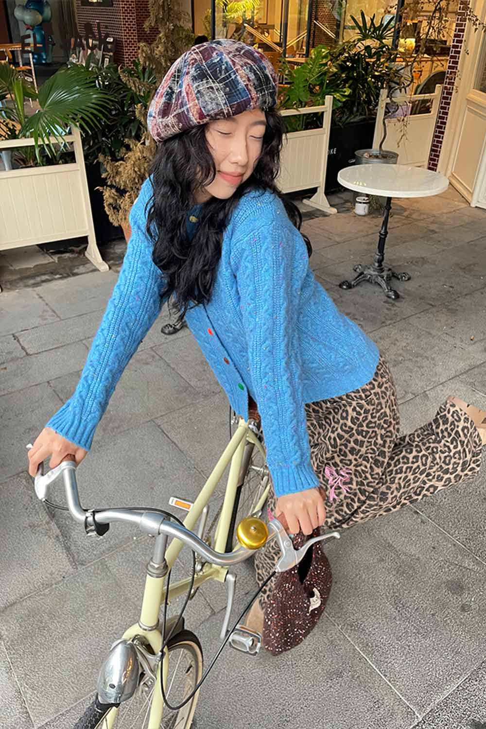 Woman wearing a color blocked knitted beret with a blue cardigan and leopard print pants, posing on a bicycle