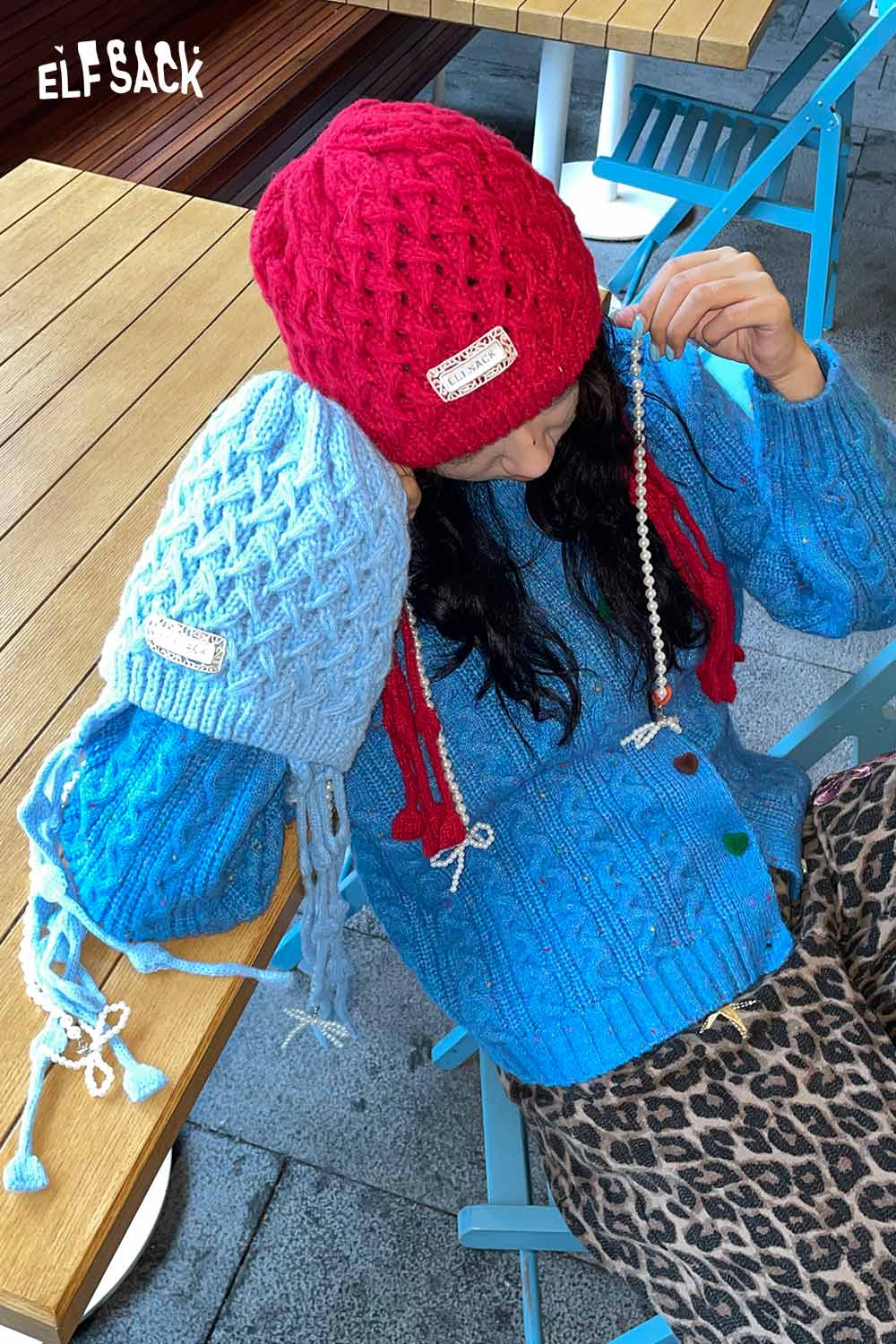 Woman wearing a blue crocheted tassel ear-flap hat alongside a red one for stylish winter warmth