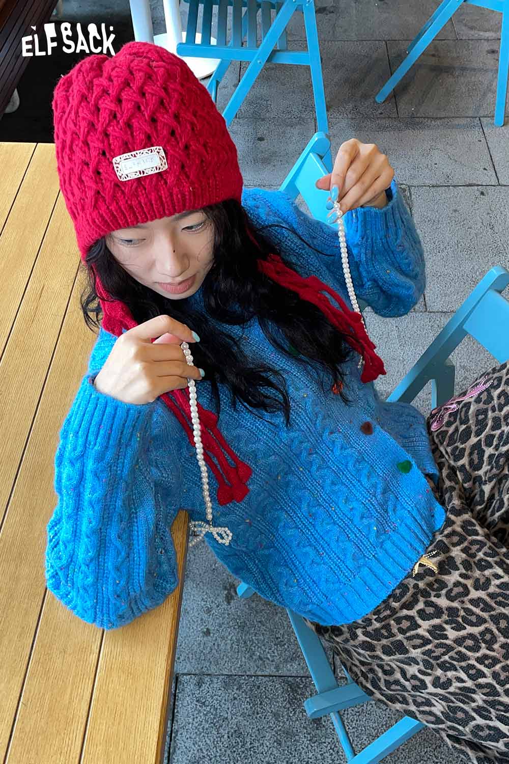 ELFSACK crocheted tassel ear-flap hat in red worn by a woman with a blue sweater and leopard print pants