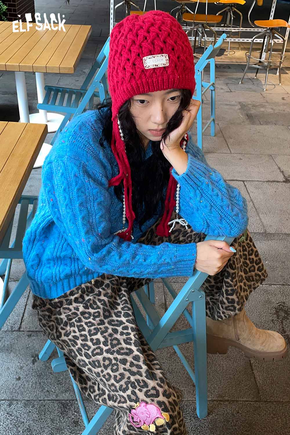 Woman wearing a bright red crocheted tassel ear-flap hat paired with a blue sweater and patterned pants