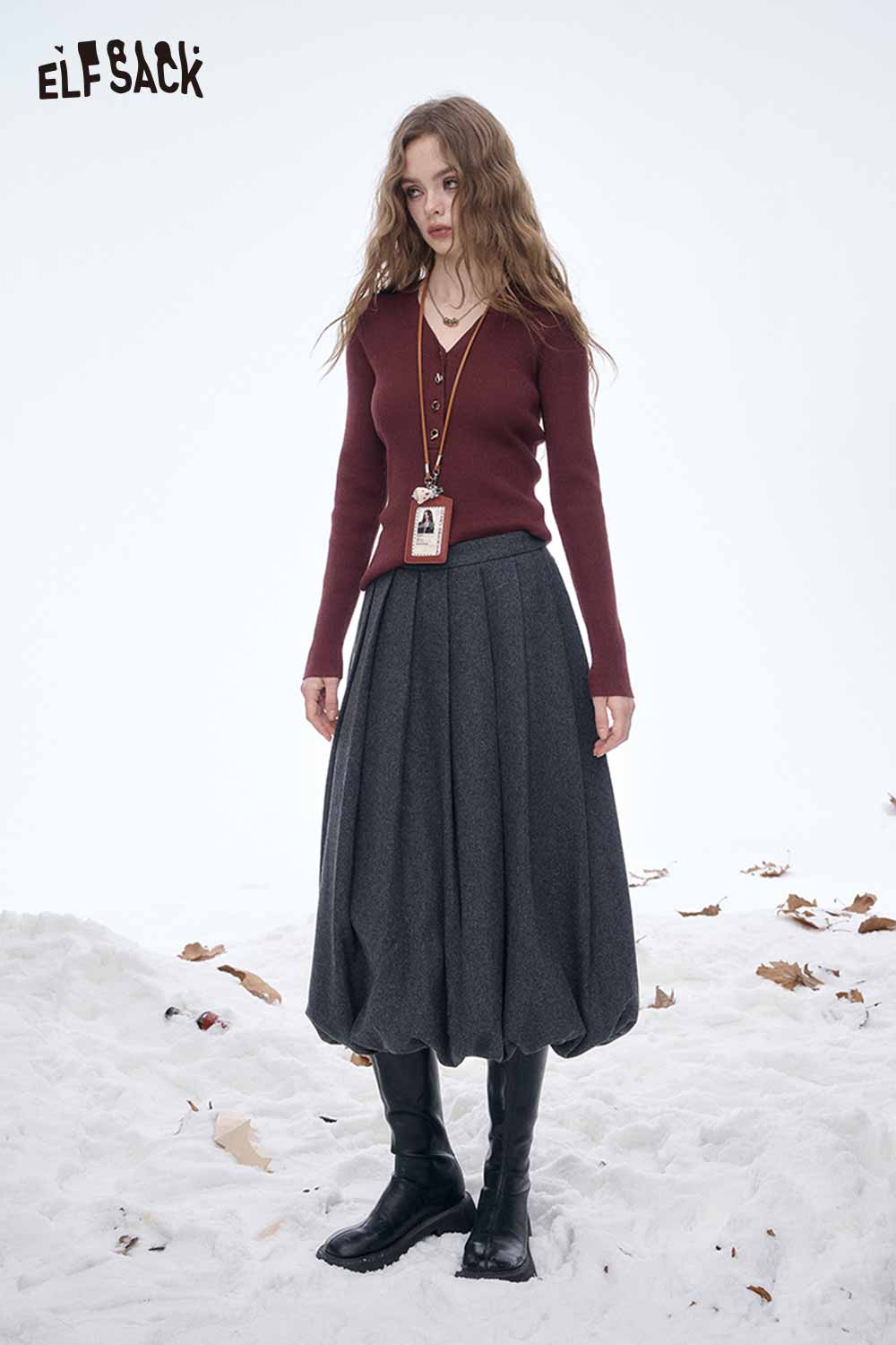 Model wearing a dark gray pleated half skirt paired with a burgundy sweater and boots in a snowy setting