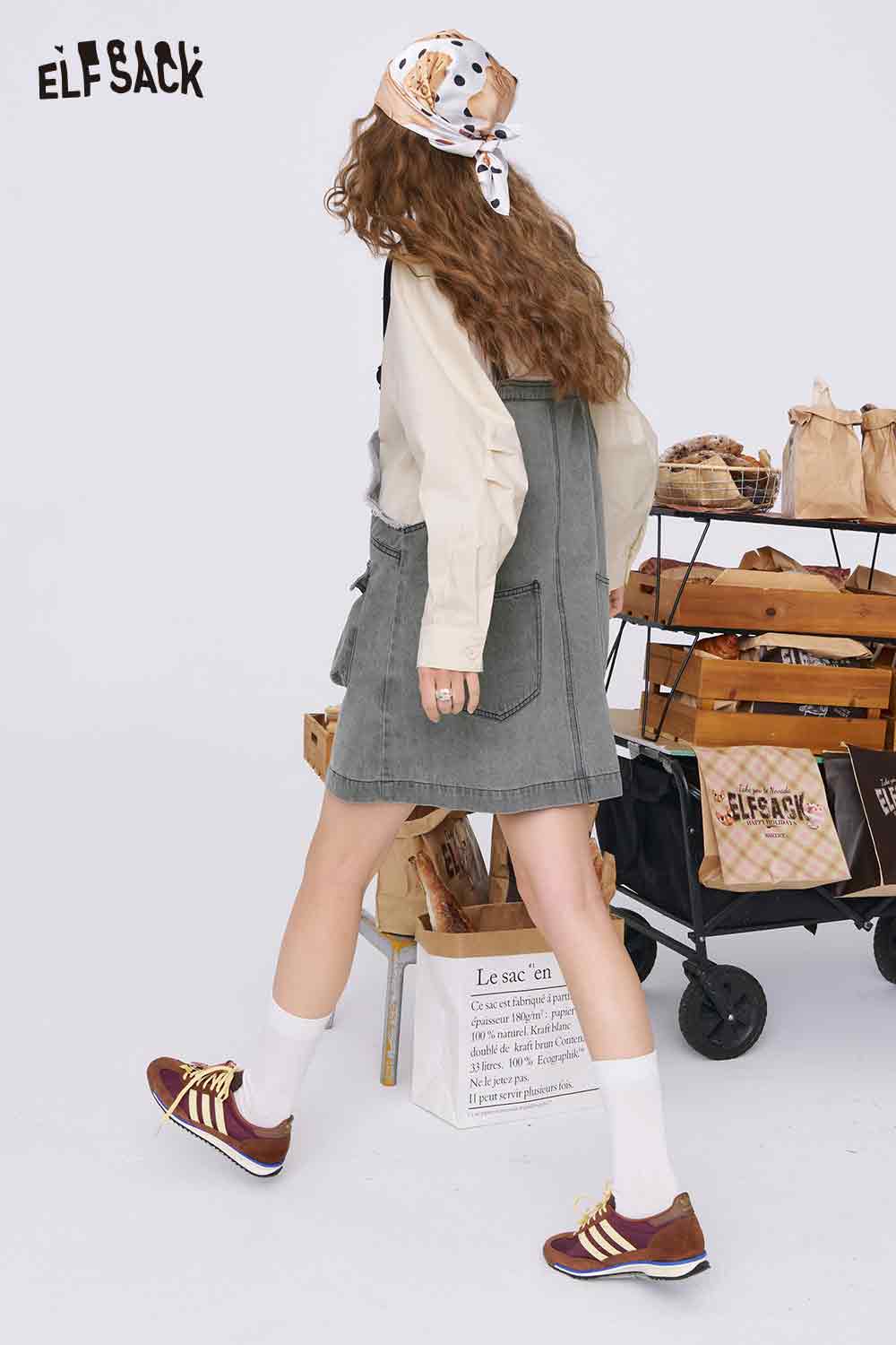 ELFSACK model wearing denim color-block midi dress pinafore with side pockets over cream blouse and brown sneakers