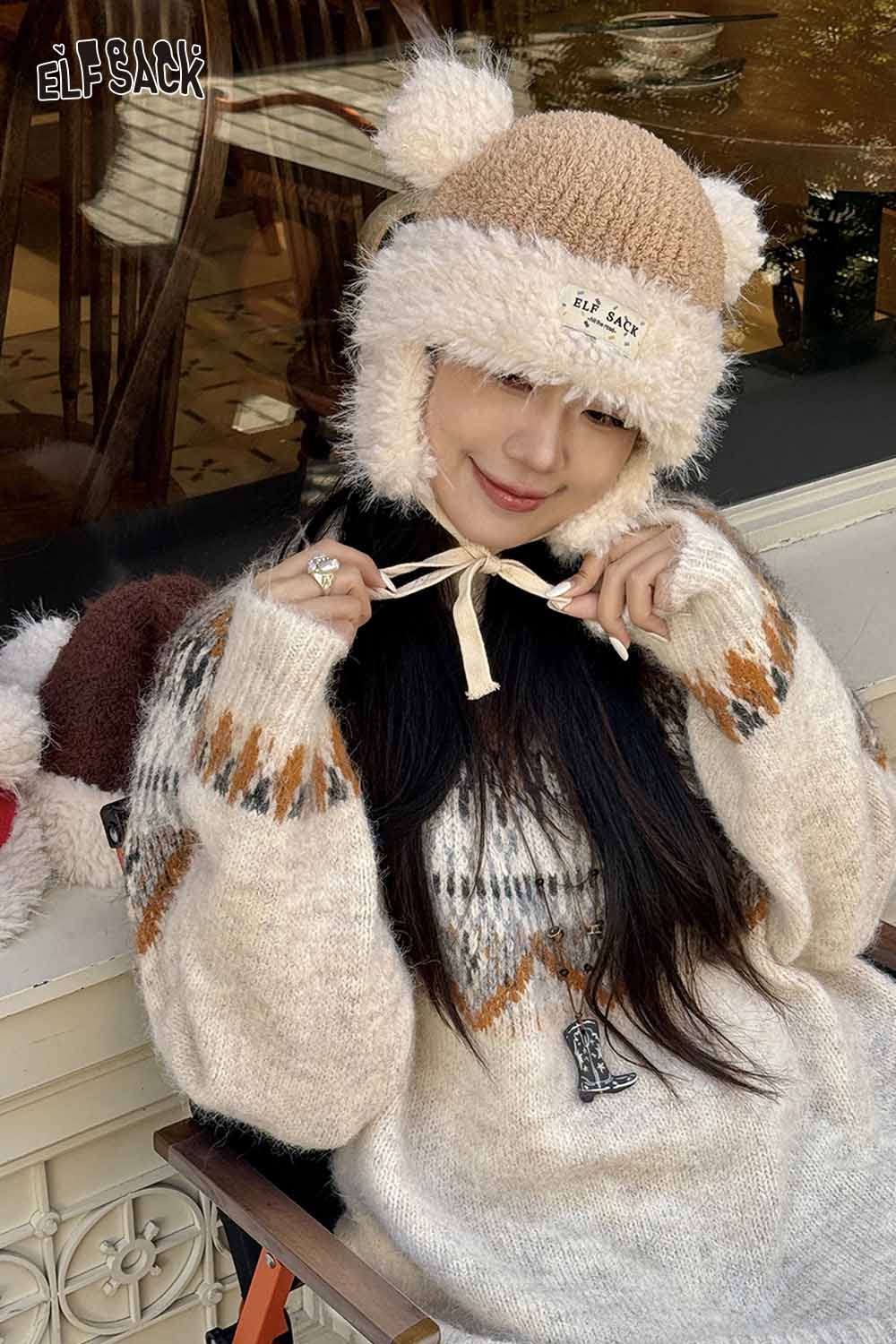 Woman wearing a cute plush ear flap hat with pom poms, styled with a cozy sweater on a patio
