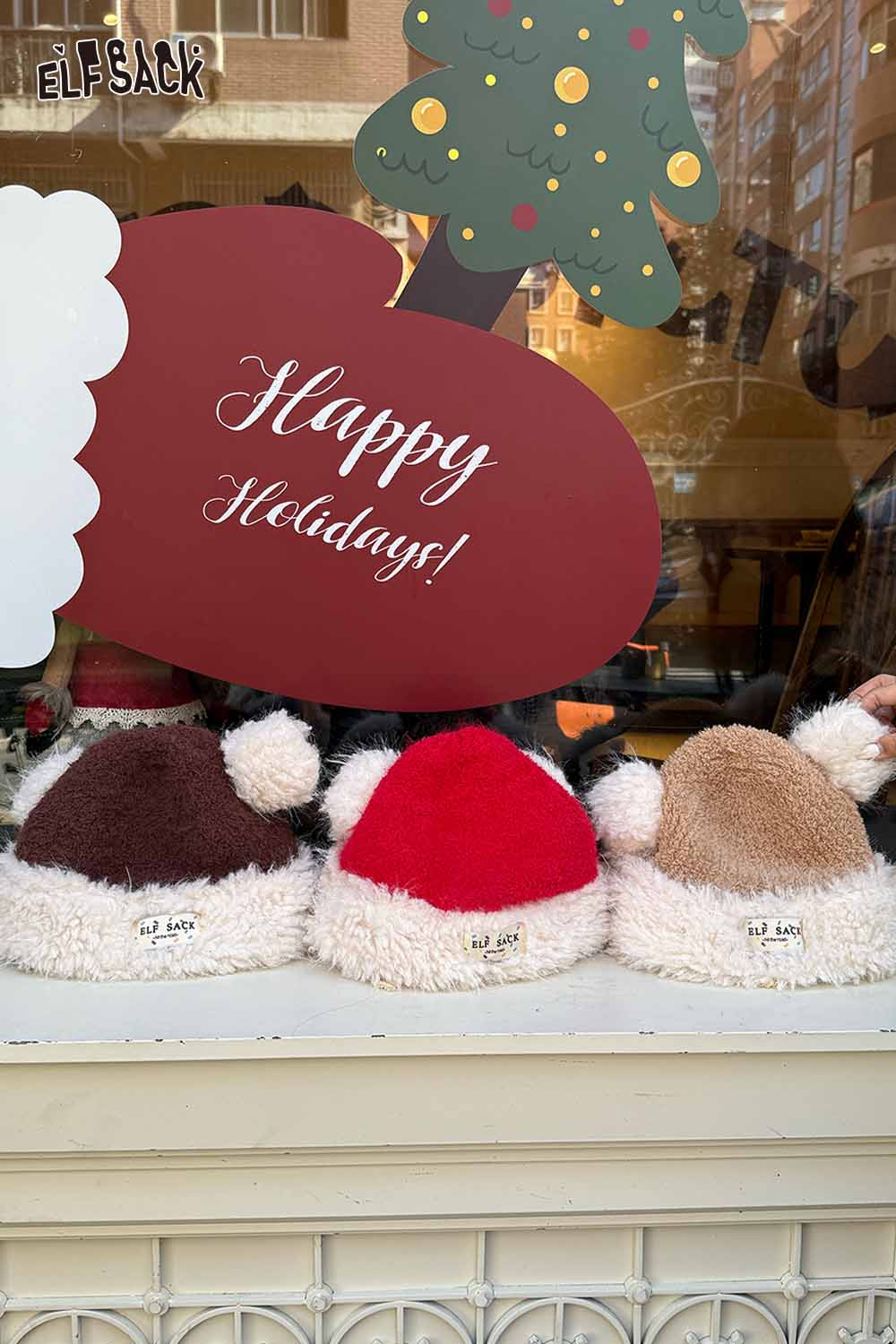 ELFSACK cute plush ear flap hat in red, brown, and beige displayed in a holiday window setting