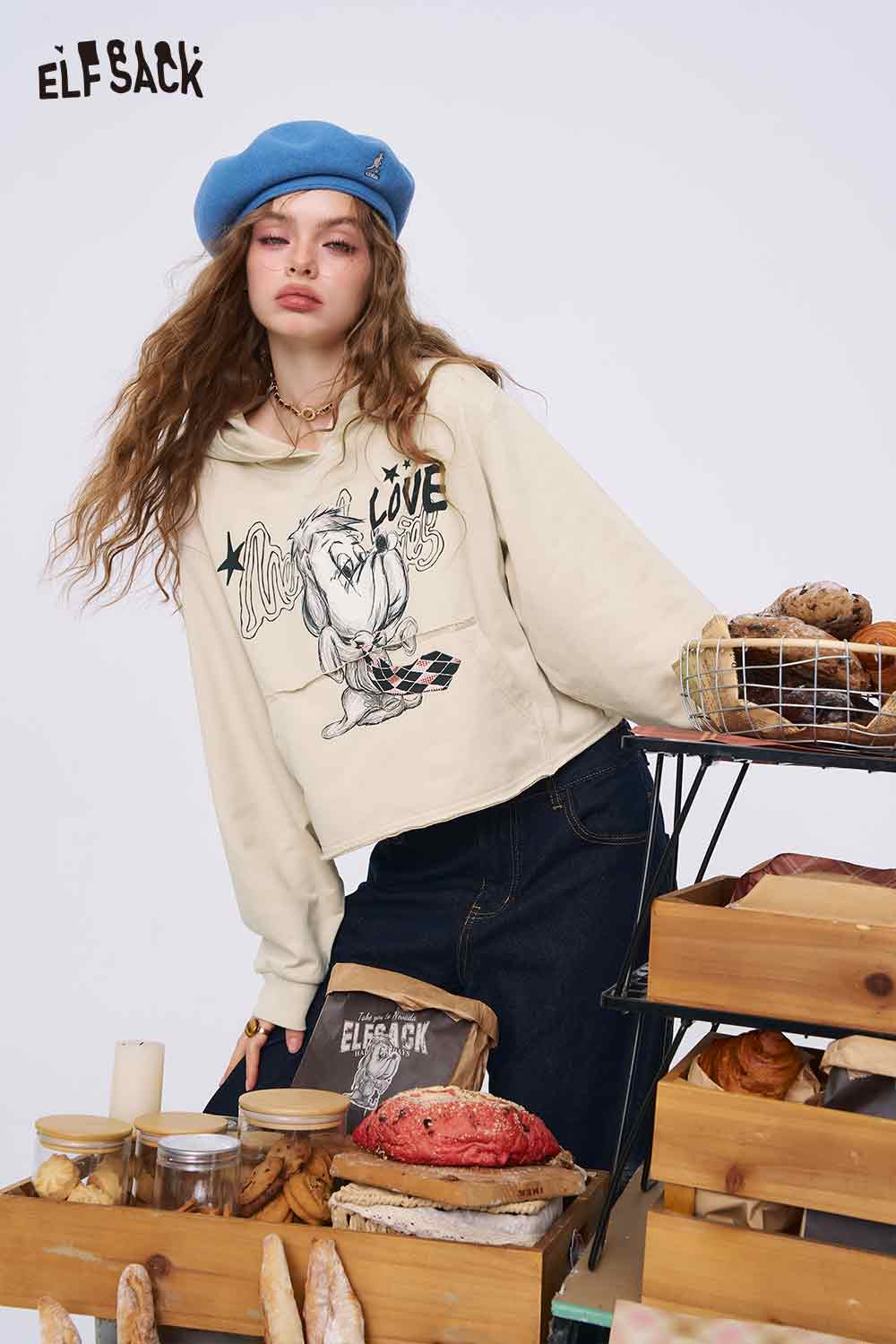 Model wearing ELFSACK beige cropped American style print hoodie for women with graphic print styled at a bakery