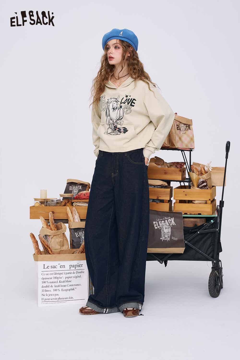 
                  
                    ELFSACK model wearing American style print hoodie for women. Cream graphic hoodie with blue beret and wide leg jeans
                  
                