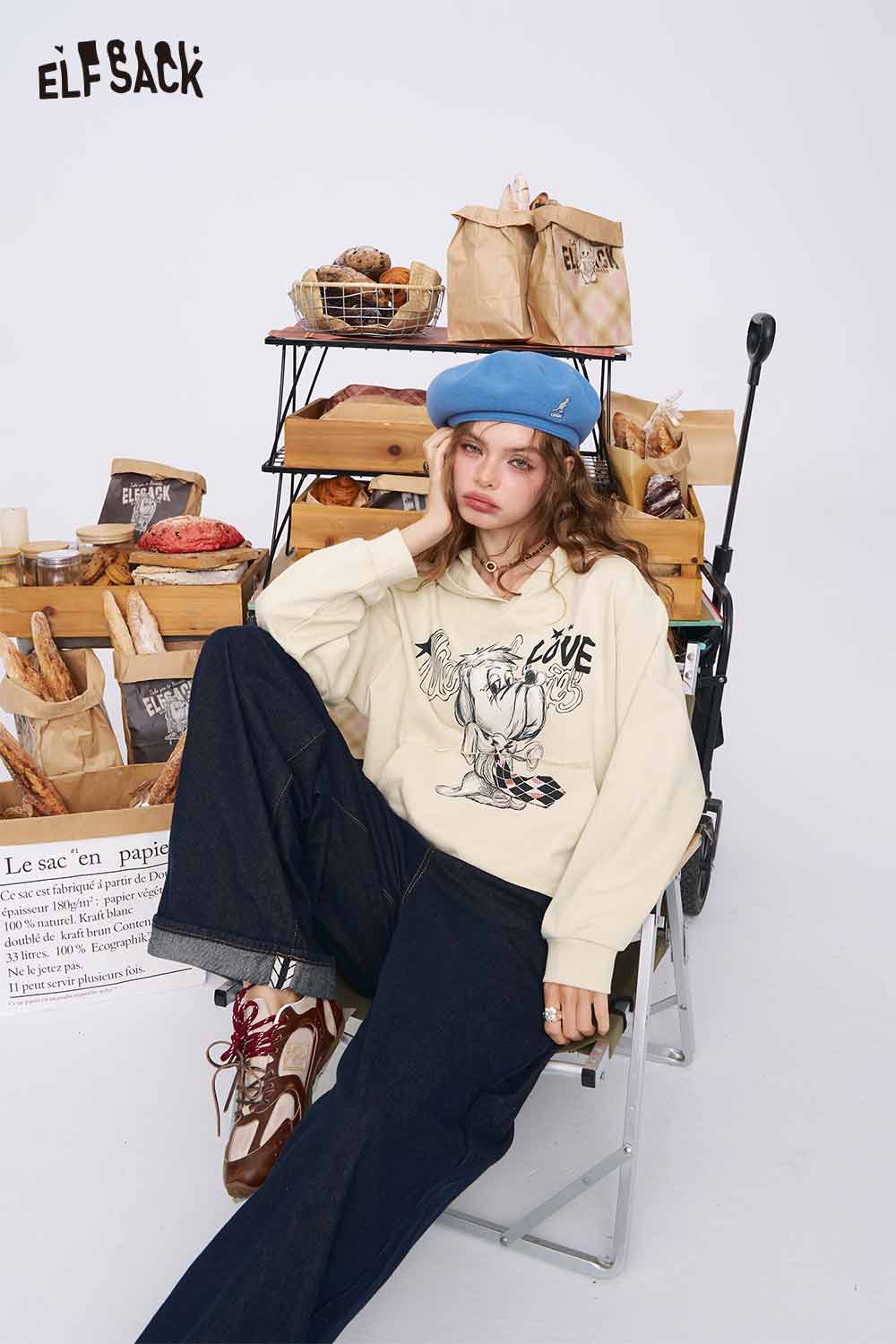 
                  
                    American style print hoodie for women. ELFSACK cream cartoon print hoodie on model with blue beret and wide leg jeans
                  
                