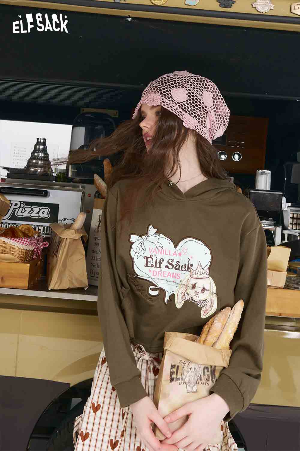 ELFSACK model wearing olive graphic American style print hoodie for women holding baguettes in a cafe setting