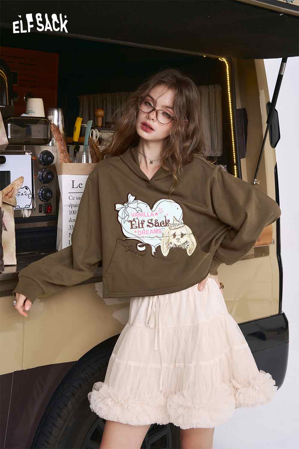 
                  
                    ELFSACK model wearing olive oversized American style print hoodie for women with white tiered tulle skirt
                  
                