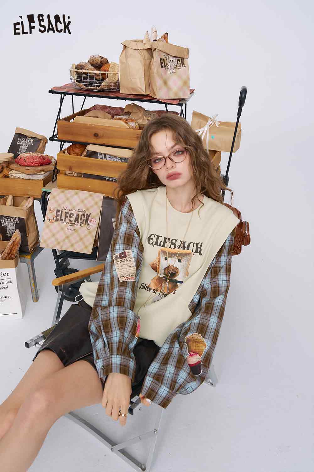 ELFSACK model wearing bread dog sweatshirt for women layered with plaid shirt seated by bakery props