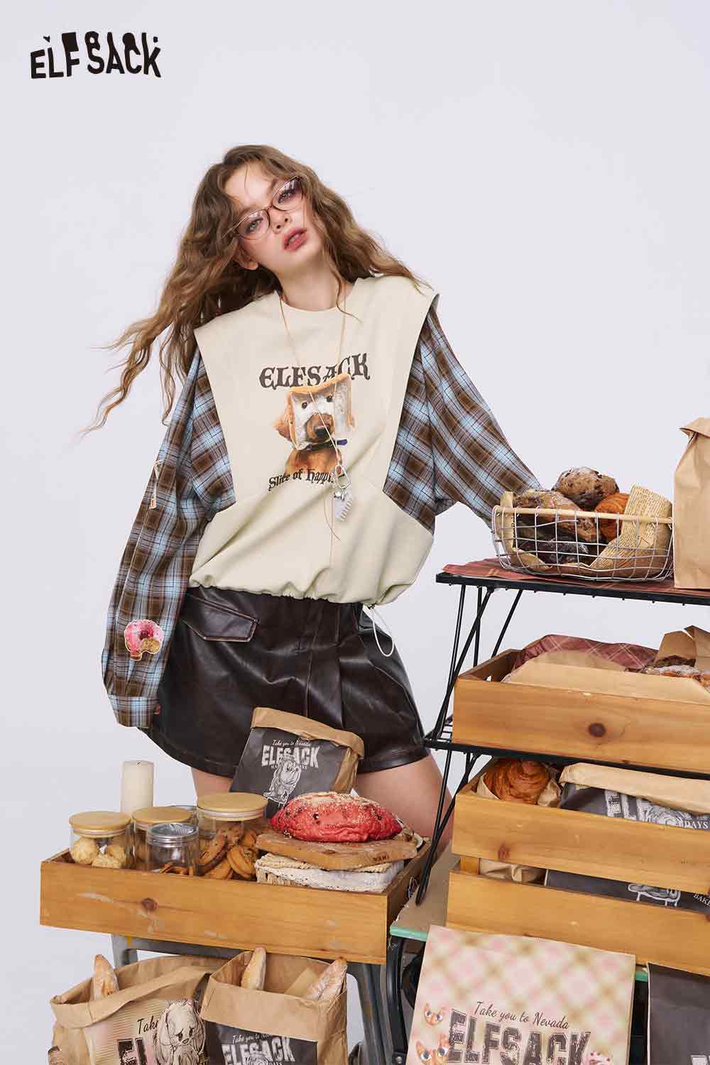 ELFSACK model wearing beige bread dog sweatshirt for women layered over plaid shirt styled with leather shorts and bakery props