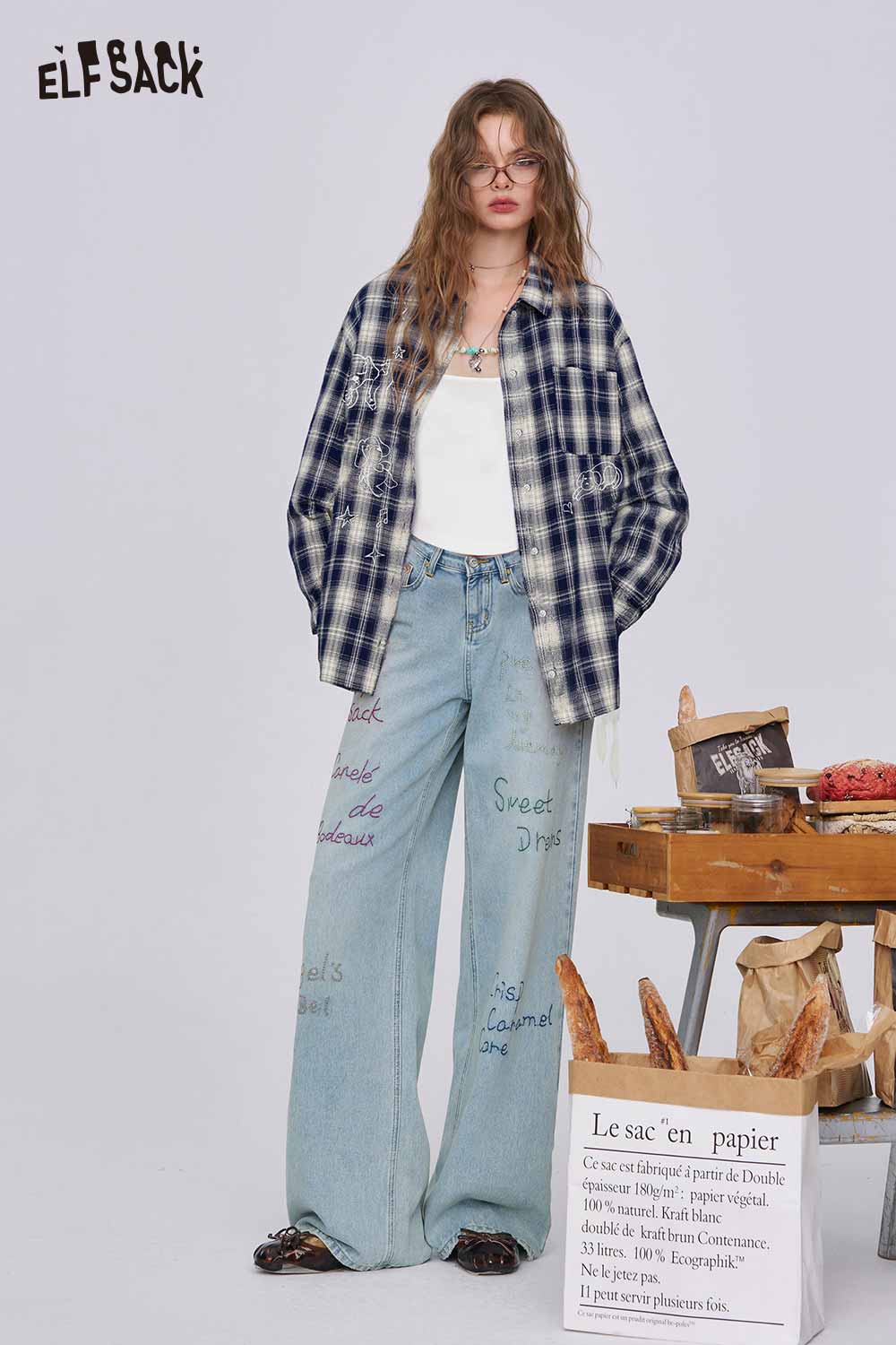 ELFSACK model wearing women's dark blue plaid graphic shirt oversized over white tank with wide leg printed jeans