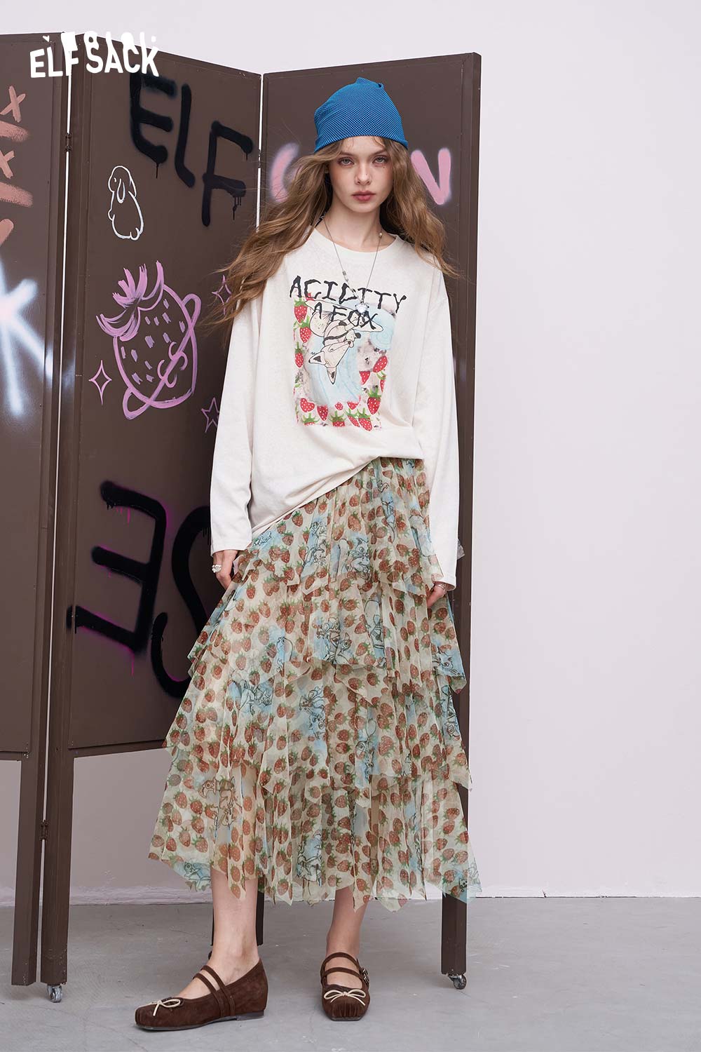 ELFSACK model wearing a strawberry print chiffon skirt in layered midi style with graphic sweatshirt and blue beanie