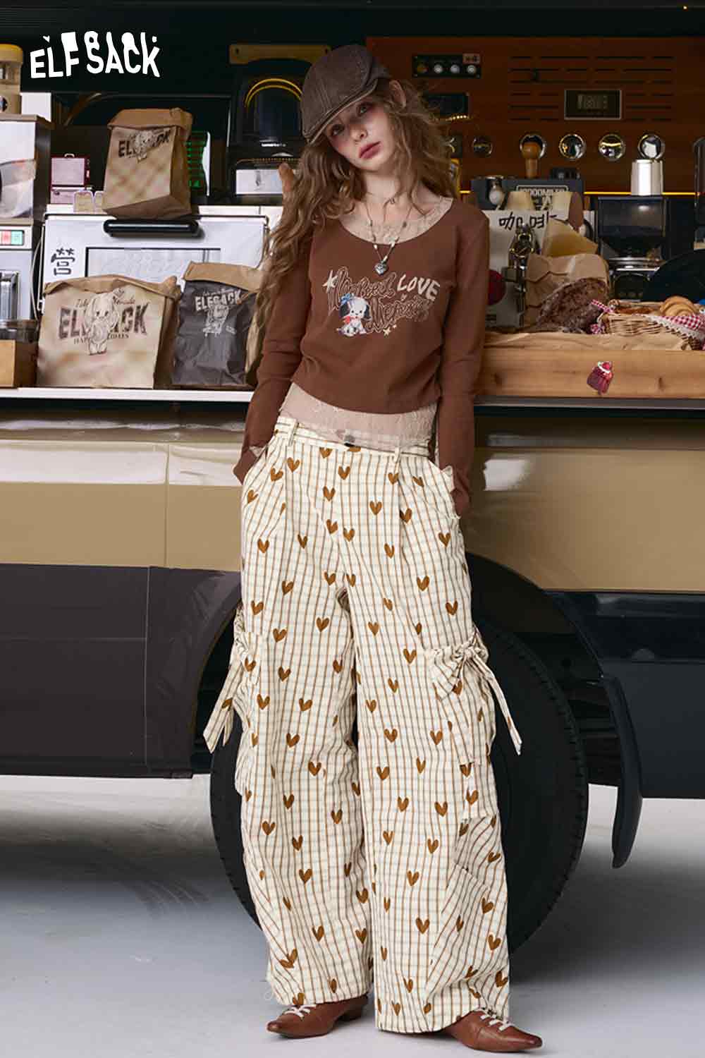 ELFSACK model wearing heart plaid wide-leg pants cream striped with brown heart print side ties paired with brown sweater and cap