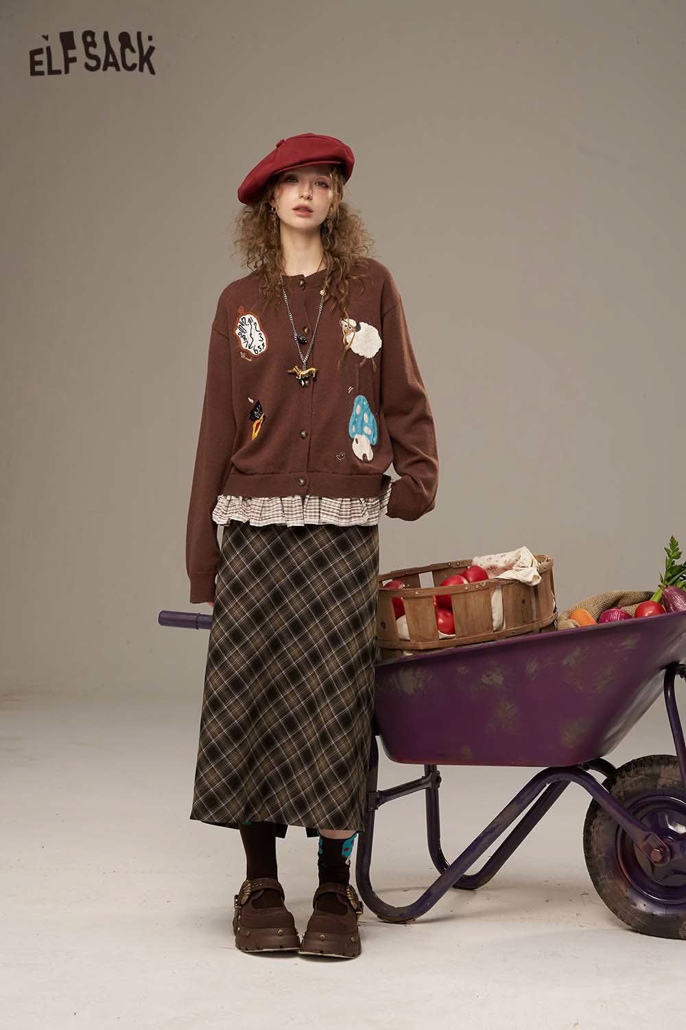 Model wearing a brown cardigan and plaid straight long skirt with a wheelbarrow in a stylish setting