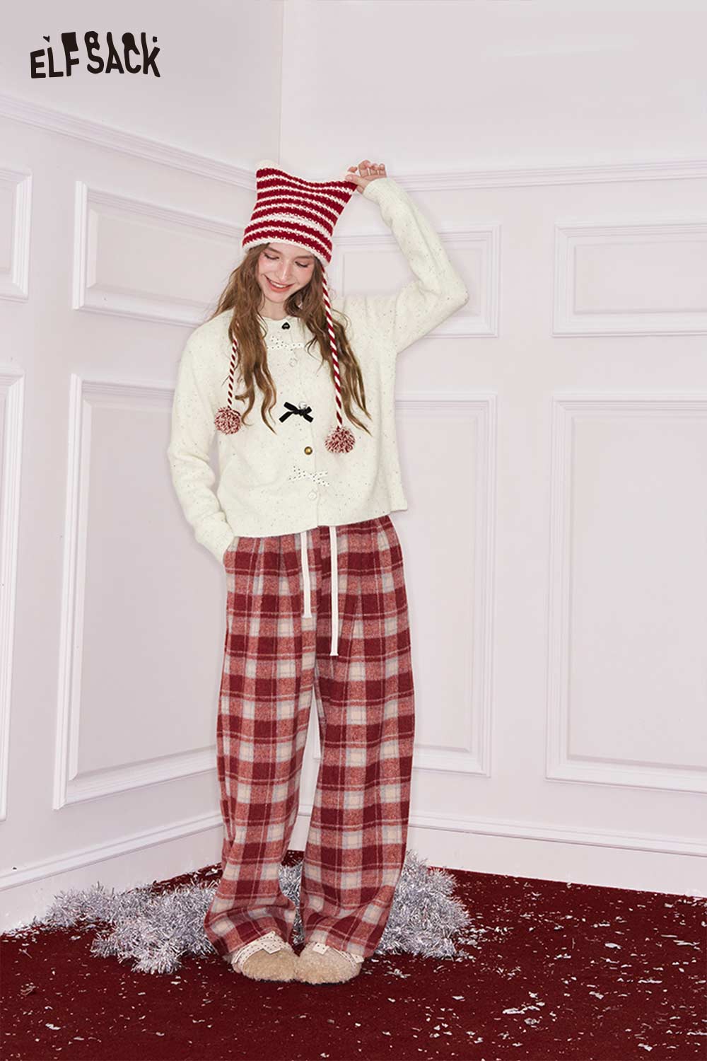 Model wearing an ELFSACK preppy 3D bow cardigan paired with plaid pants in a cozy setting