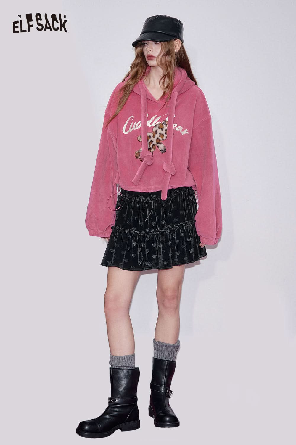 Model wearing a preppy black velvet mini skirt styled with a pink hoodie and black boots