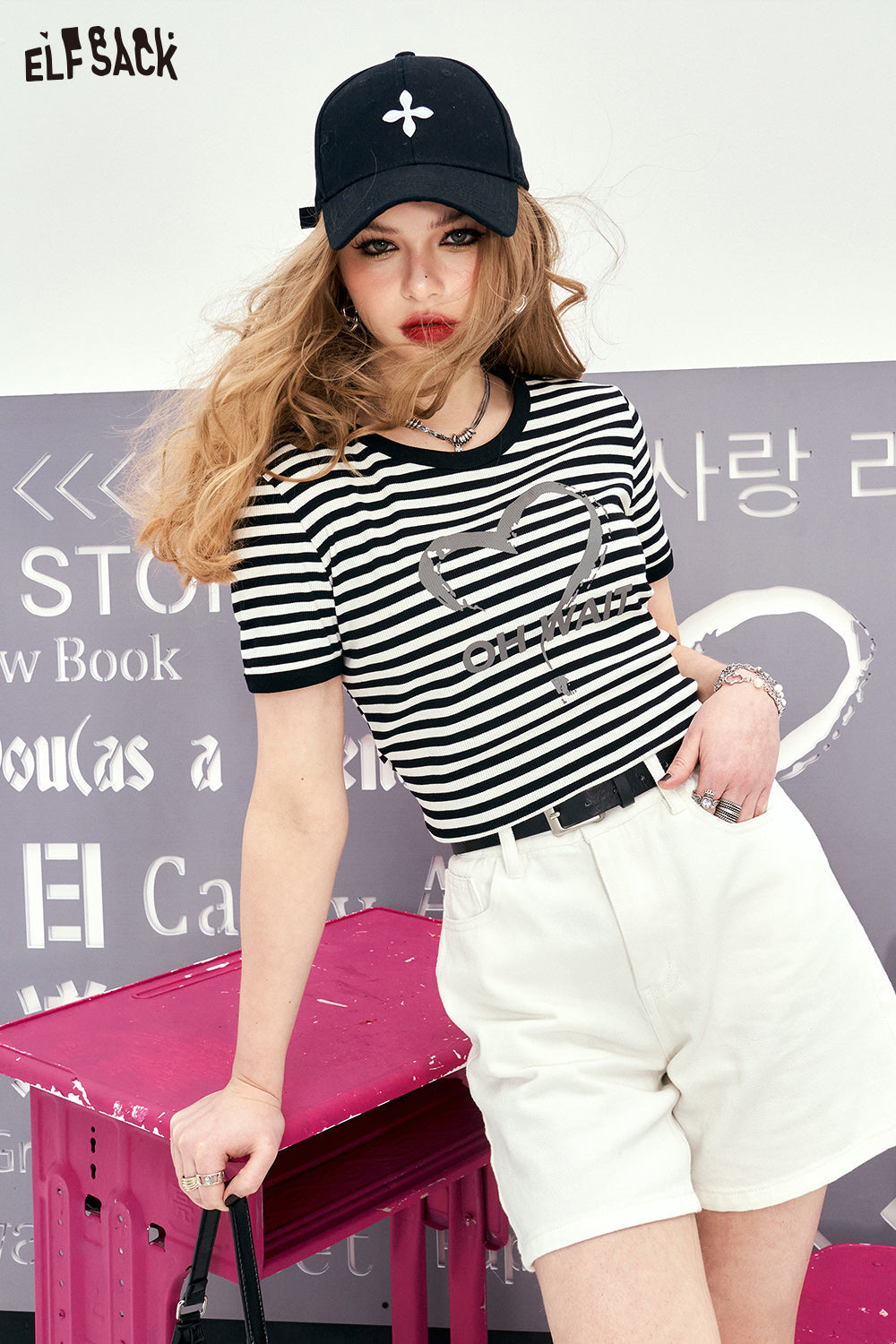 Retro black and white striped ELFSACK T-shirt with heart design worn by woman with white shorts and black cap