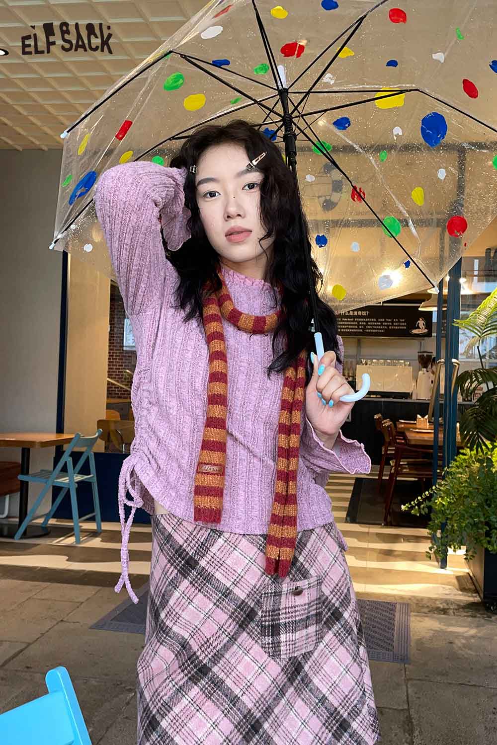 Trendy woman wearing a pink sweater and soft striped scarf for women with an umbrella in a colorful cafe setting
