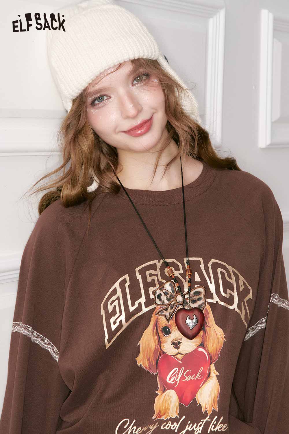 
                  
                    ELFSACK model wearing a brown graphic sweatshirt with a vintage heart pendant necklace
                  
                