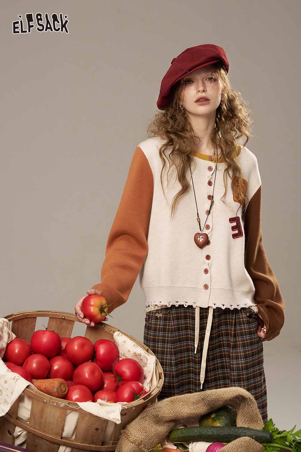 ELFSACK women's knitted jacket in cream and burnt orange worn with a plaid skirt and beret while holding apples