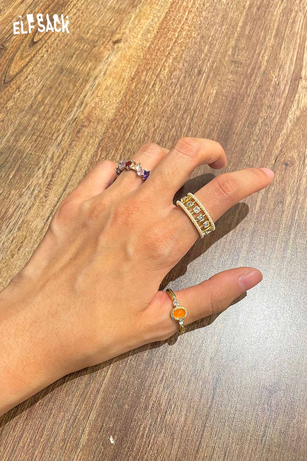 ELFSACK zirconia gemstone copper ring displayed on a hand with multiple rings under warm lighting